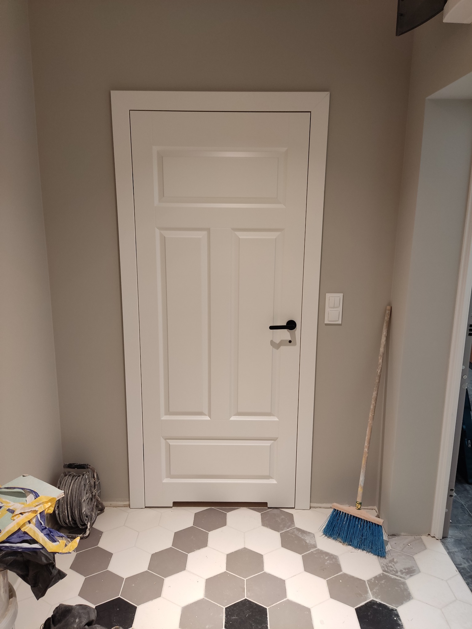 Białe drzwi wewnętrzne z czarną klamką, świeżo po montażu, widoczne narzędzia i akcesoria malarskie na podłodze z heksagonalnych płytek w odcieniach szarości.