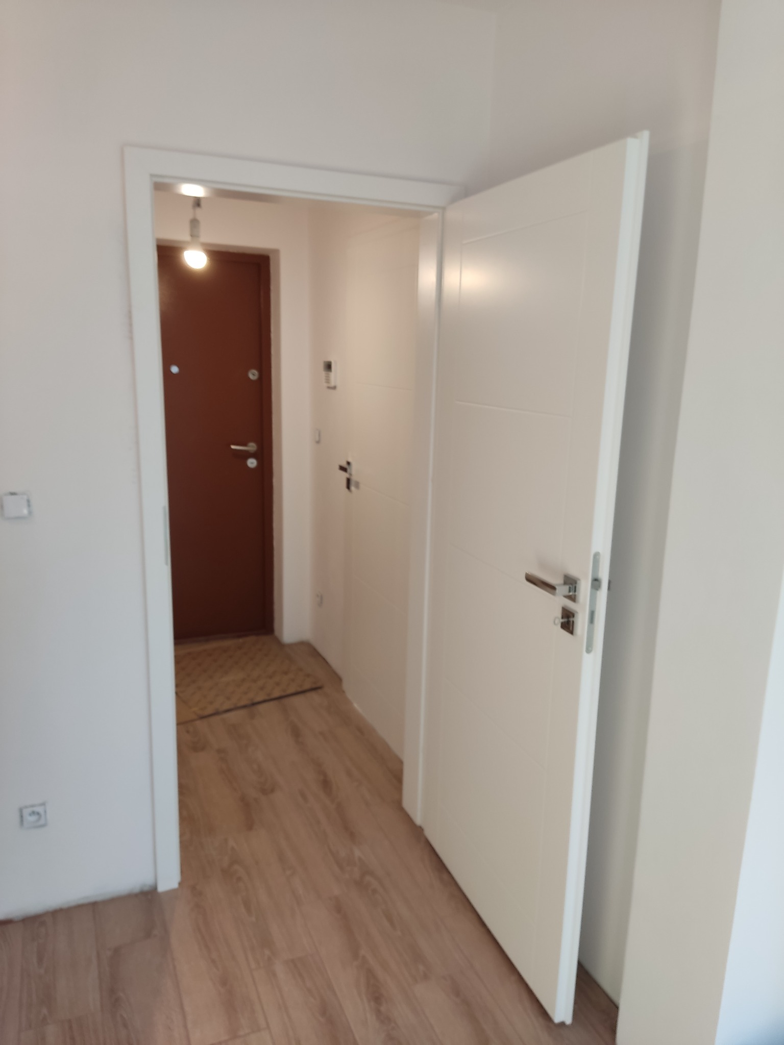 Otwarta biała, wewnętrzna płycinowa drzwi z chromowaną klamką, widok na przedpokój z brązowymi drzwiami wejściowymi i drewnianą podłogą.