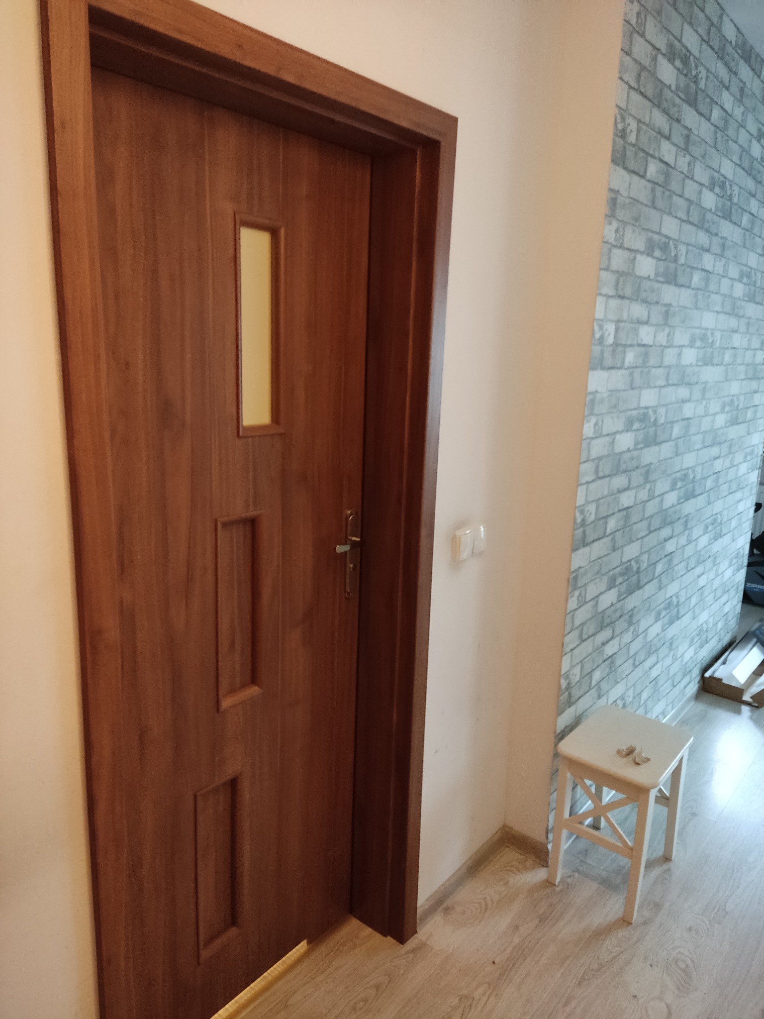 Zamknięte, drewniane drzwi wewnętrzne z trzema prostokątnymi wgłębieniami i małym okienkiem, zamontowane w pokoju z szarą ścianą z cegły i białym taboretem.