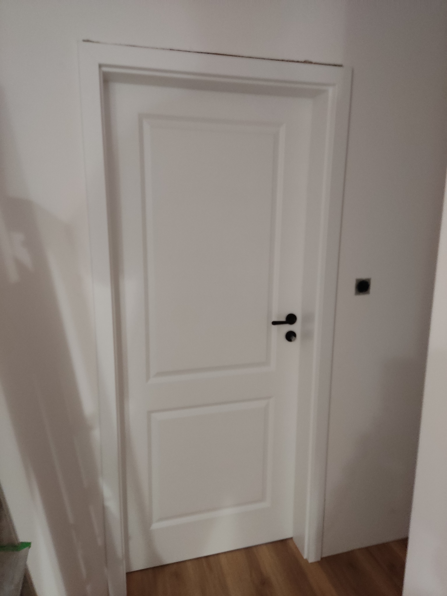 Zamknięte, białe drzwi wewnętrzne z czarną klamką i dwoma panelami, widoczny fragment podłogi z paneli i gniazdko elektryczne na ścianie.