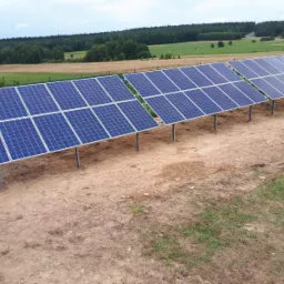 Dwie rzędy paneli słonecznych zamontowane na metalowych słupkach na tle pola i lasu, widoczne z perspektywy lekko z góry.