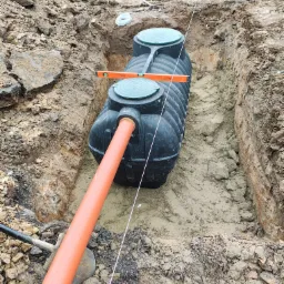 Czarny zbiornik z rurą w wykopie, poziomica na górze. Widok z góry na instalację w ziemi. Wykopane ściany ziemne, lina pionowa, narzędzia w tle.