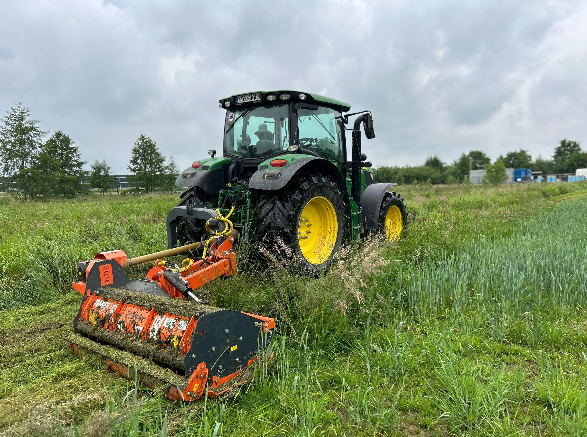 Zielony traktor John Deere z pomarańczową kosiarką rotacyjną na polu, widok z tyłu, pochmurne niebo w tle. Kosiarka w trakcie koszenia trawy.
