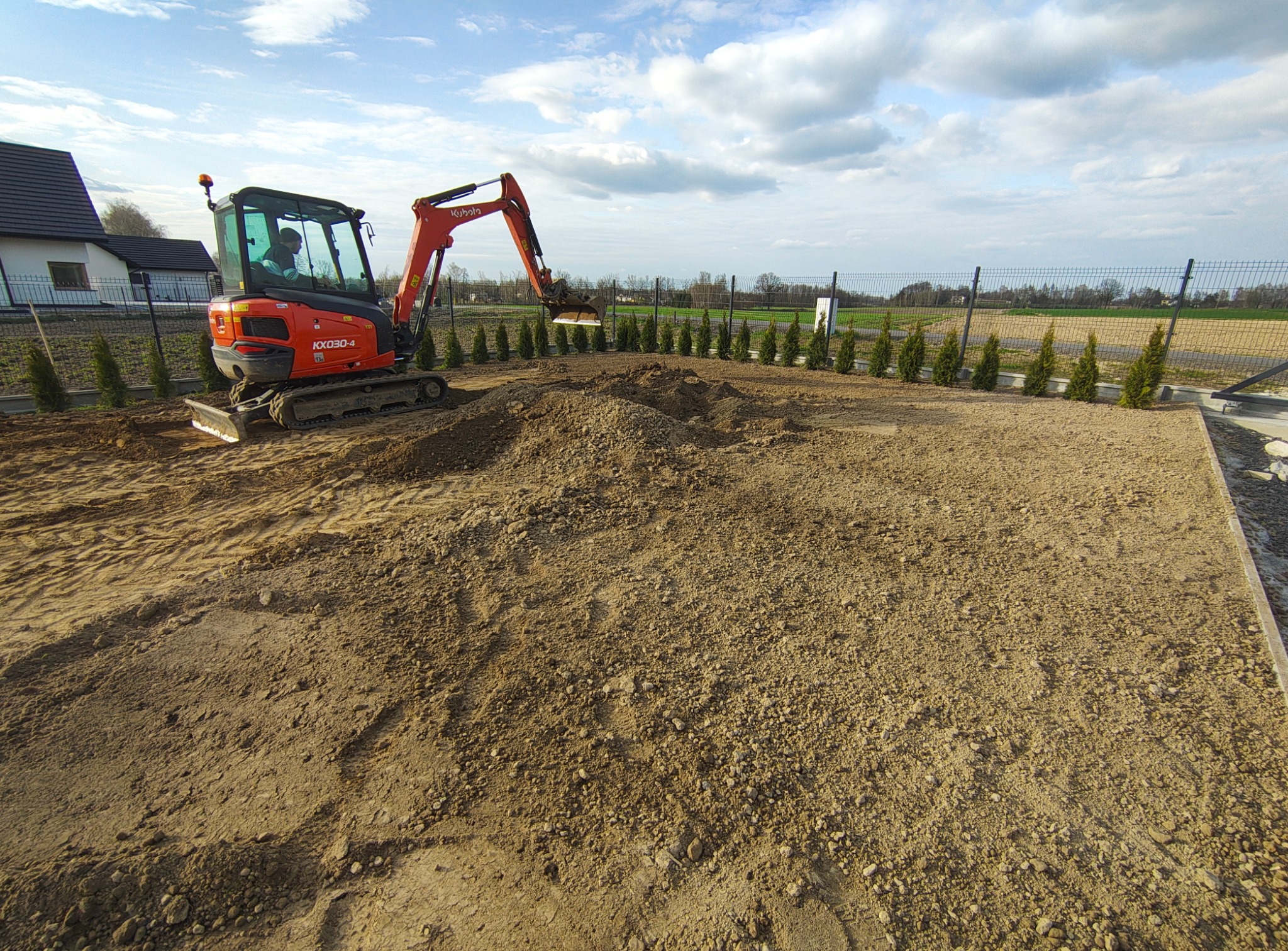 Pomarańczowa mini koparka Kubota KX030-4 niweluje teren działki z młodymi tujami w rzędzie, widok z góry na wyrównywaną ziemię, błękitne niebo z chmurami.
