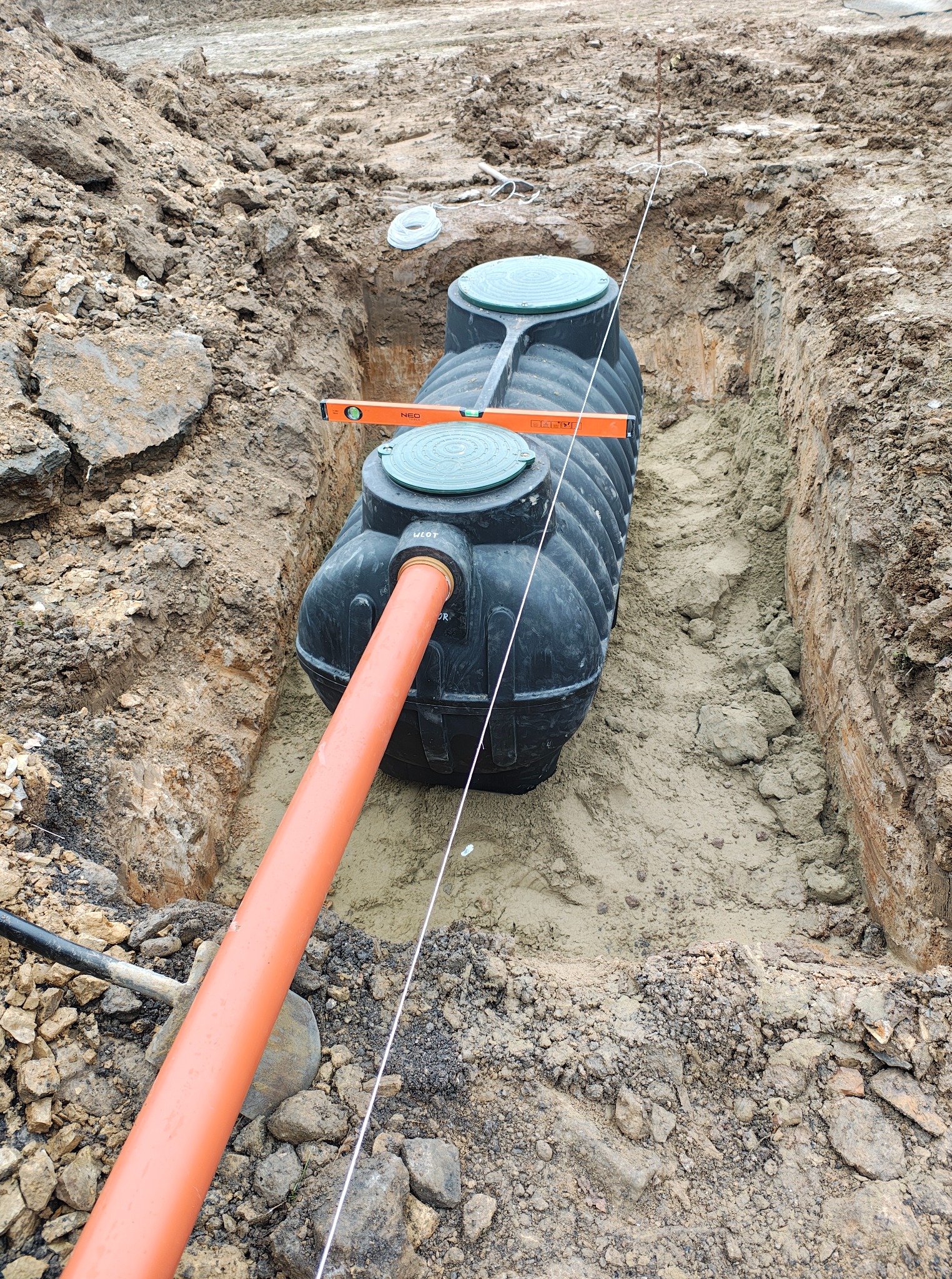 Czarny zbiornik z rurą w wykopie, poziomica na górze. Widok z góry na instalację w ziemi. Wykopane ściany ziemne, lina pionowa, narzędzia w tle.