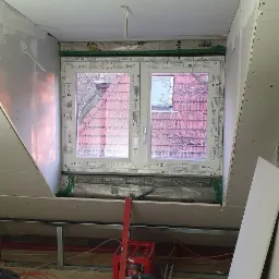 Poddasze w trakcie adaptacji: widoczne okno dachowe, ocieplenie folią paroizolacyjną, płyty kartonowo-gipsowe w trakcie montażu na skosach, metalowy profil, narzędzia i wiadro z gładzią szpachlową...