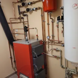 Czerwony piec na paliwo stałe z czarnym kominem, obok biały zasobnik ciepłej wody użytkowej i czerwony zbiornik wyr&oacute;wnawczy, połączone miedzianymi rurami z zaworami i manometrami.