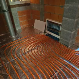 Montaż ogrzewania podłogowego z pomarańczowych rur na folii z nadrukiem, widoczny rozdzielacz w białej obudowie i surowe ściany z cegły dziurawki oraz betonowych bloczków.