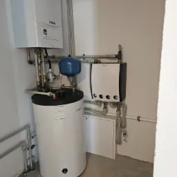 Kompaktowa kotłownia w narożniku pomieszczenia: wiszący piec gazowy Vaillant, biały zasobnik ciepłej wody z niebieskim naczyniem wzbiorczym oraz dodatkowy biały moduł grzewczy z perforowaną obudową...