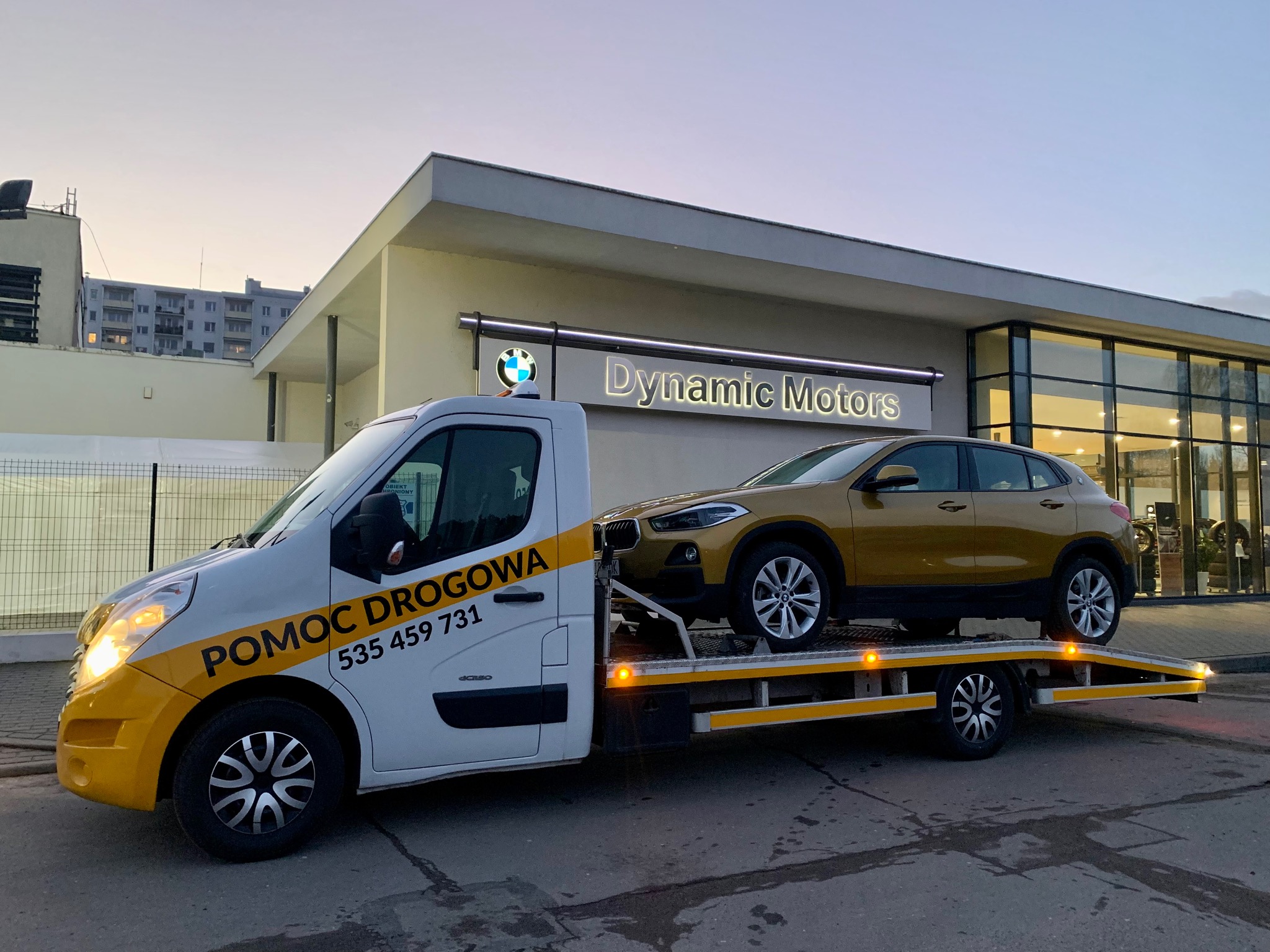 Złoty samochód BMW X2 na platformie lawety z napisem 'POMOC DROGOWA 535 459 731' na tle salonu samochodowego 'Dynamic Motors' o zmierzchu.