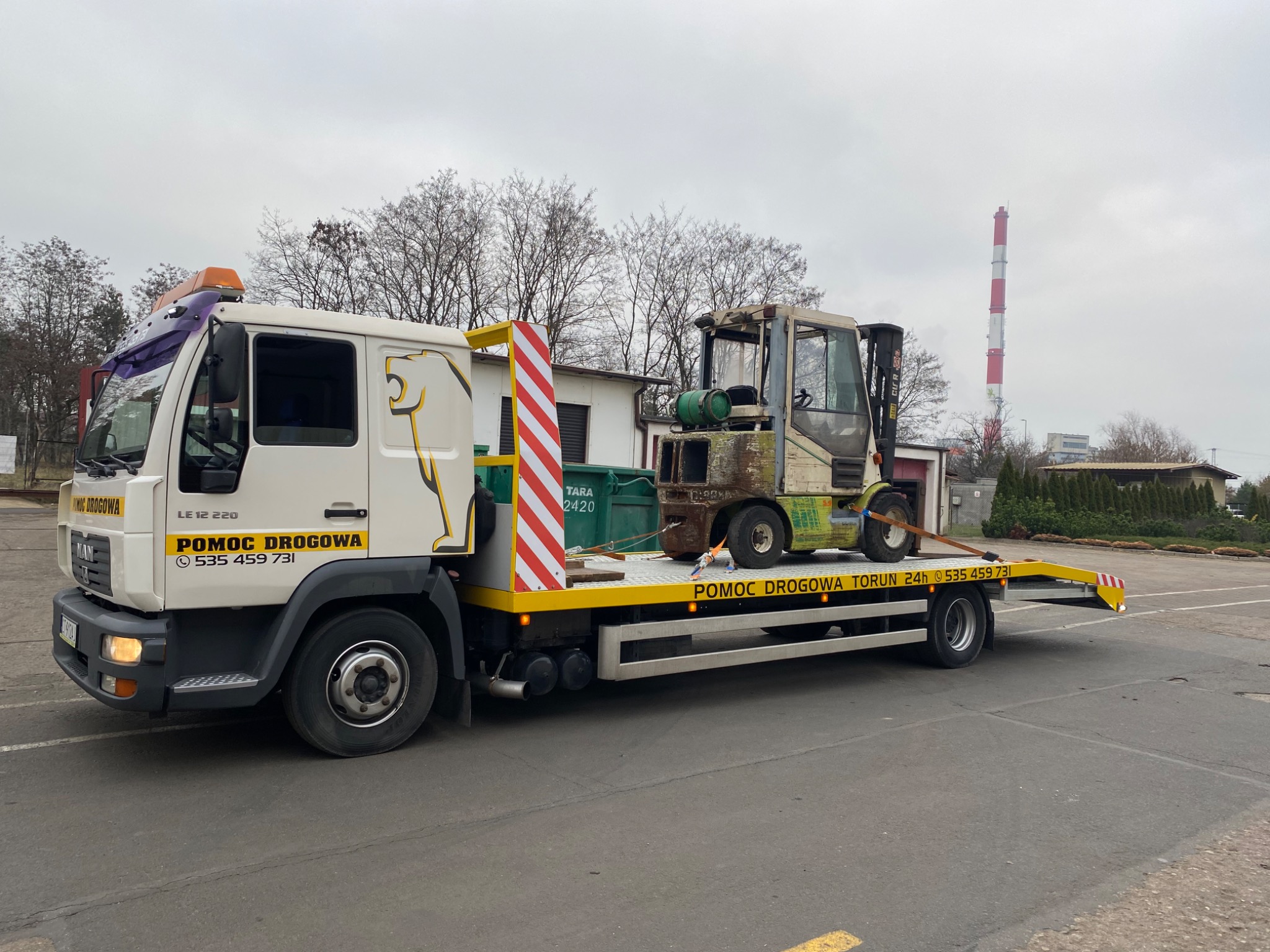 Biała laweta z żółtymi elementami i logo lwa przewozi zniszczony wózek widłowy na platformie. W tle budynki i wysoki komin.