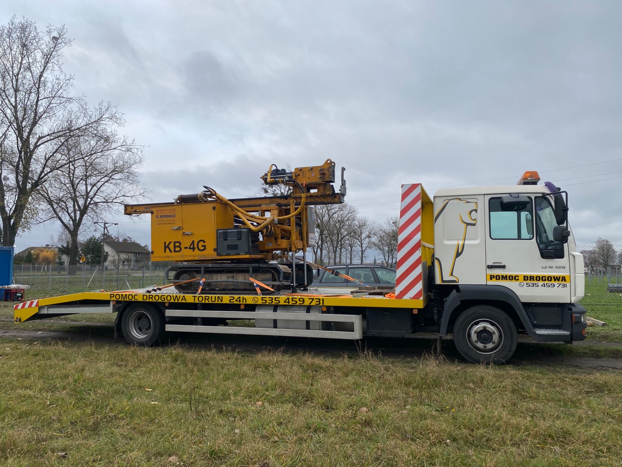 Żółta wiertnica KB-4G zabezpieczona pasami na platformie pojazdu pomocy drogowej z napisem 'POMOC DROGOWA TORUN 24h' na tle pochmurnego nieba i trawiastym terenie.