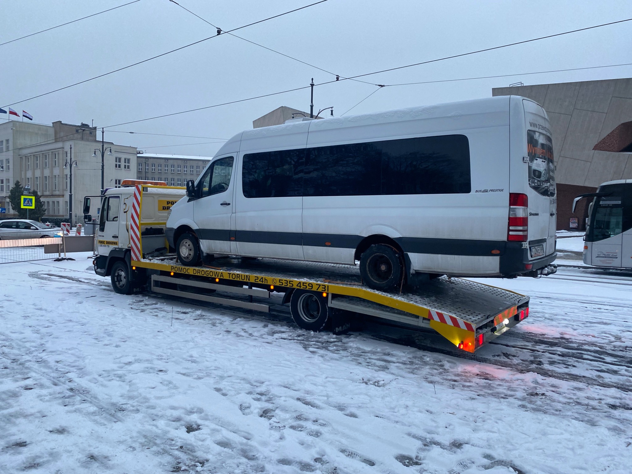 Biały bus na lawecie pomocy drogowej w zimowej scenerii miejskiej Torunia, widoczny numer telefonu alarmowego, budynki w tle.