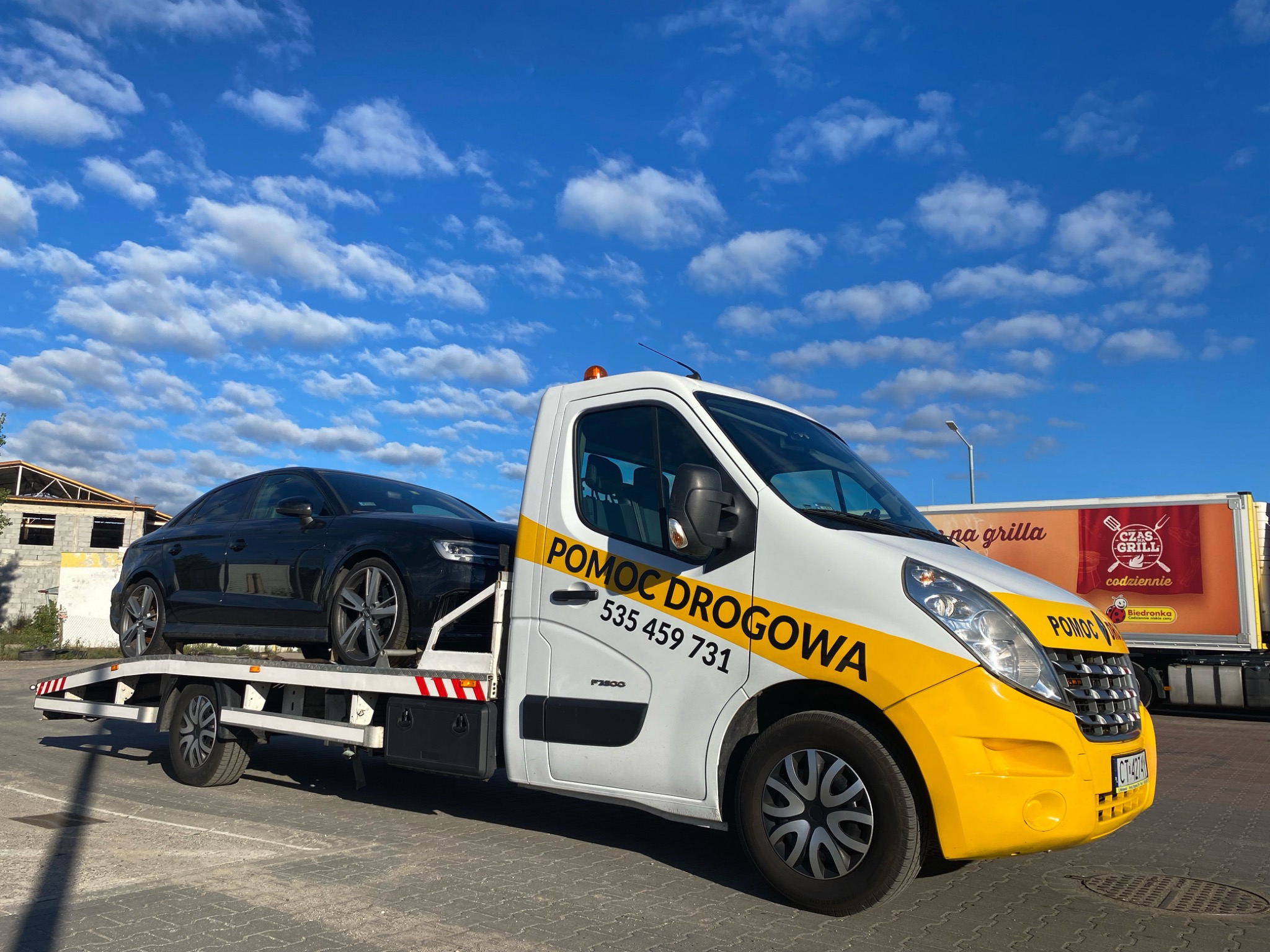 Czarny samochód osobowy marki Audi transportowany na białej lawecie z żółtymi elementami i napisem 'Pomoc Drogowa' na tle błękitnego nieba z chmurami.