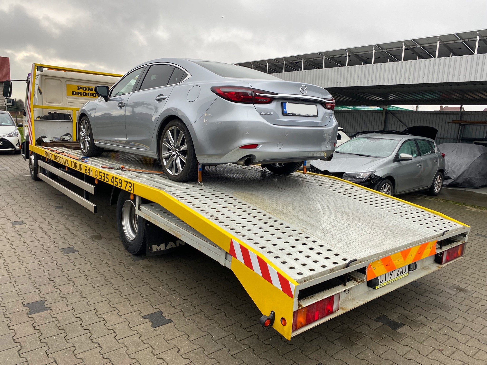 Srebrny samochód osobowy Mazda 6 zabezpieczony pasami na żółtej platformie pomocy drogowej MAN, widok z tyłu, na placu z kostki brukowej w pochmurny dzień.