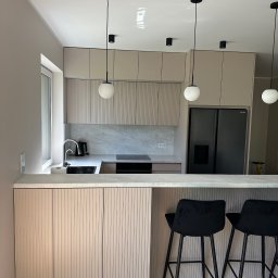 TOP MEBLE STEFANI DAMIAN STEFANI - Nowoczesna kuchnia z beżowymi frontami szafek z ryflowanym wzorem, czarnymi lampami wiszącymi i marmurowym blatem. Dwa czarne hokery przy wyspie.