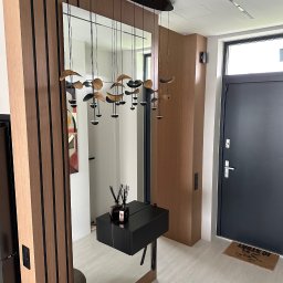 TOP MEBLE STEFANI DAMIAN STEFANI - Nowoczesny przedpokój z lustrem i drewnianą zabudową. Uwagę przyciąga designerska lampa wisząca i minimalistyczna konsola z czarnym blatem.