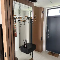 Nowoczesny przedpokój z lustrem i drewnianą zabudową. Uwagę przyciąga designerska lampa wisząca i minimalistyczna konsola z czarnym blatem.