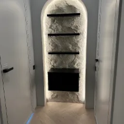 Narożna wnęka z oświetleniem LED, teksturą kamienia i czarnymi półkami, otoczona białymi drzwiami z czarnymi klamkami. Jasny parkiet ułożony w jodełkę.