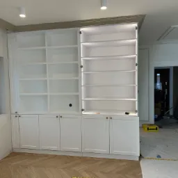 Biała zabudowa meblowa z półkami i szafkami, jedna szafka z oświetleniem LED, parkiet w jodełkę. Widoczne ślady prac wykończeniowych.