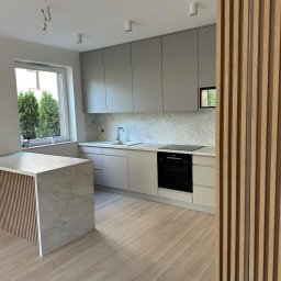 TOP MEBLE STEFANI DAMIAN STEFANI - Jasna kuchnia z wyspą, fronty szafek w odcieniach szarości i beżu, marmurowy blat i backsplash. Drewniane lamele w tle dodają ciepła.