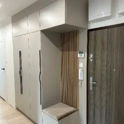 Nowoczesna szafa wnękowa z siedziskiem i pionowymi lamelami w przedpokoju, minimalistyczny design z czarnymi uchwytami i drewnianą okleiną drzwi.