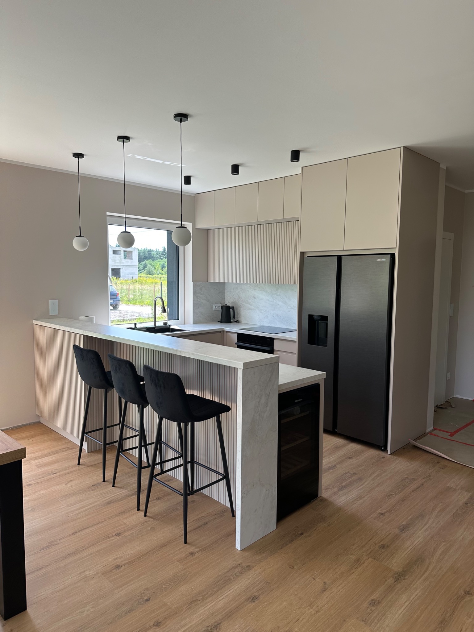 Jasna kuchnia z wyspą i trzema czarnymi hokerami. Uwagę przykuwa marmurowy blat wyspy i minimalistyczne lampy wiszące nad nią. Nowoczesne wykończenie i sprzęt AGD.