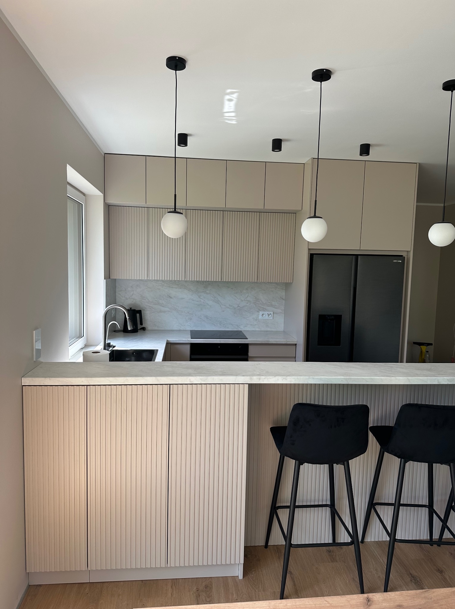 Nowoczesna kuchnia z beżowymi frontami szafek z ryflowanym wzorem, czarnymi lampami wiszącymi i marmurowym blatem. Dwa czarne hokery przy wyspie.