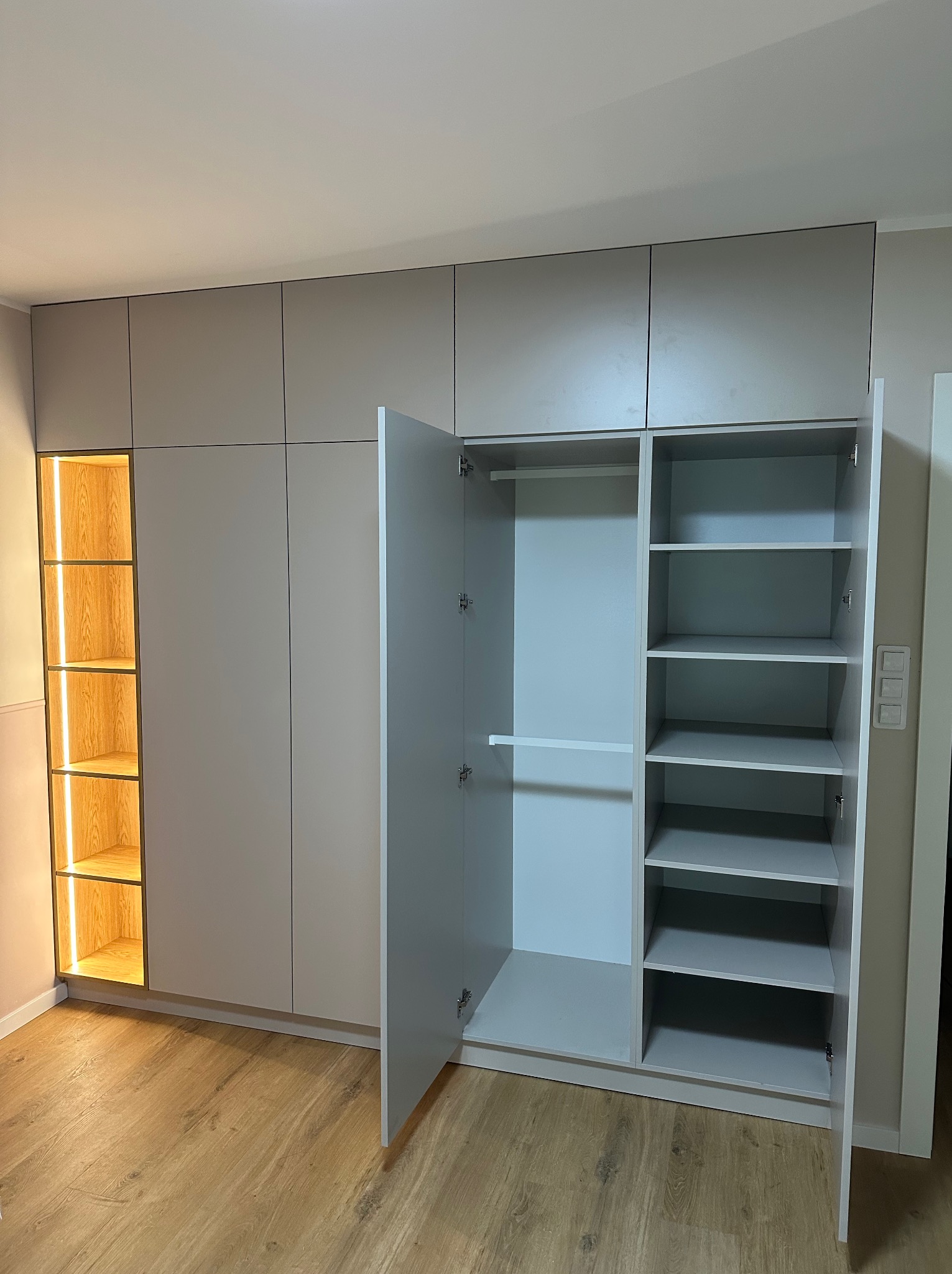 Szafa wnękowa w odcieniach szarości z otwartymi drzwiami, ukazująca półki i wieszaki. Po lewej stronie podświetlane półki z drewnianym wykończeniem.