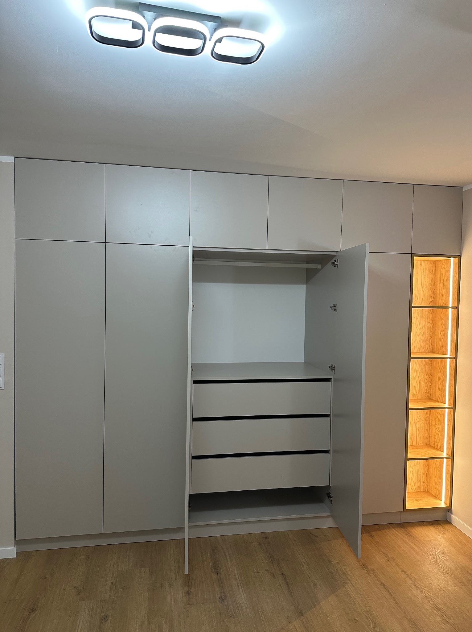 Jasnoszara szafa na wymiar z otwartymi drzwiami, widoczne szuflady i półki z podświetleniem LED. Minimalistyczny design, drewniana podłoga.