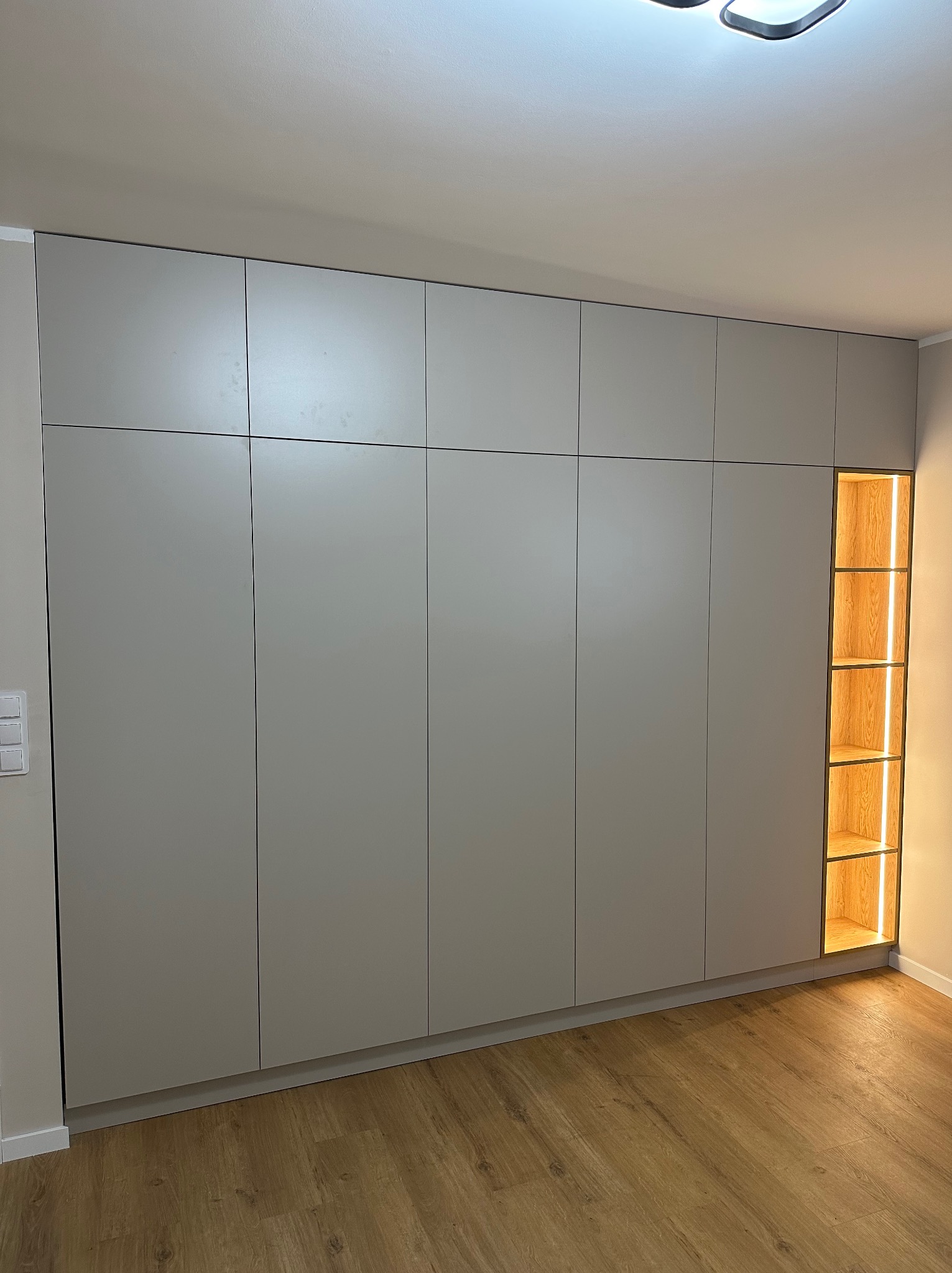 Duża, szara szafa wnękowa z oświetloną, drewnianą wnęką z półkami. Minimalistyczny design, drewniana podłoga. Nowoczesne rozwiązanie do przechowywania.