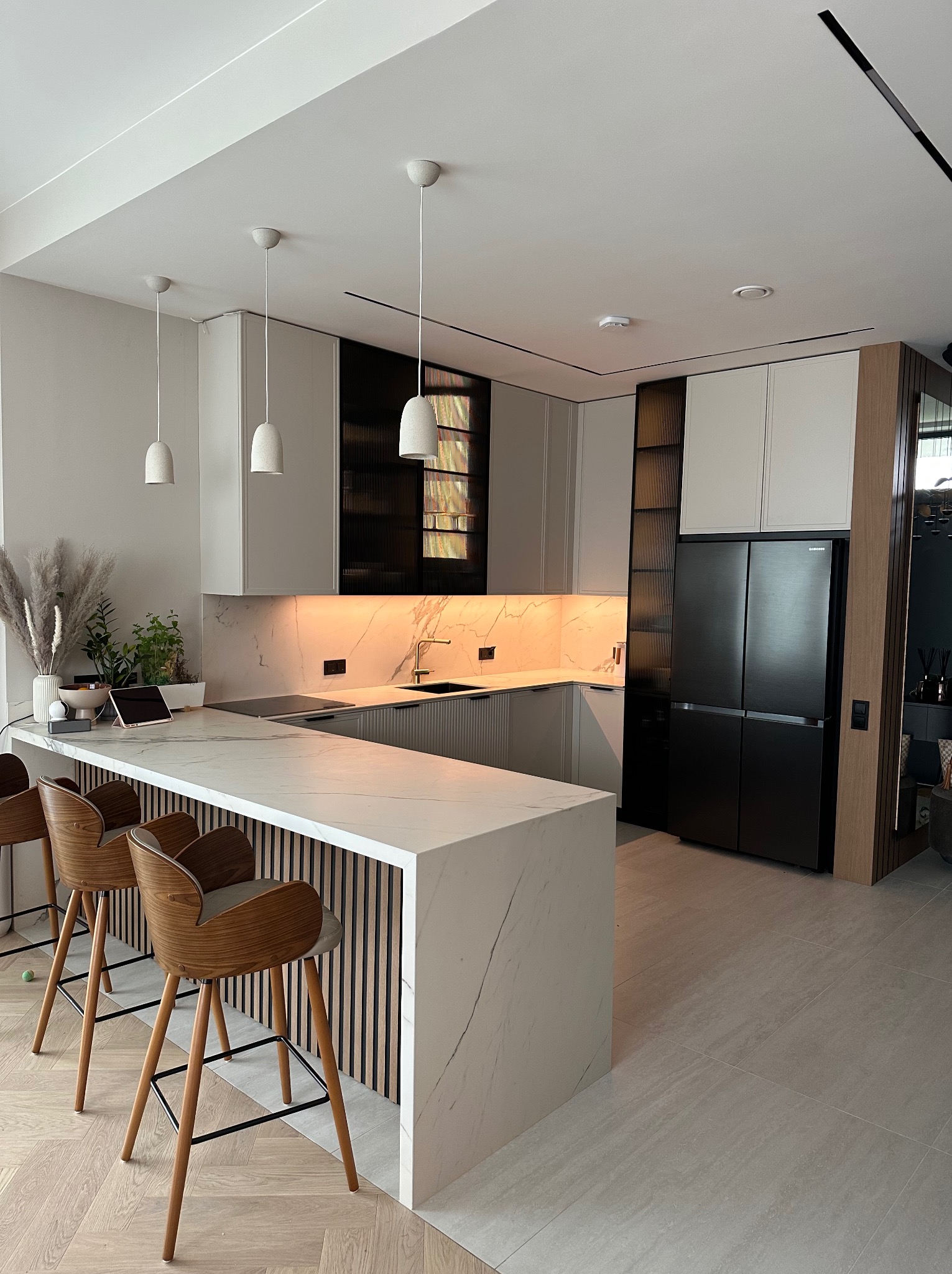Nowoczesna kuchnia z wyspą, marmurowy blat, drewniane krzesła barowe. Minimalistyczne lampy wiszące, czarna lodówka i szafki z ryflowanymi szybami.