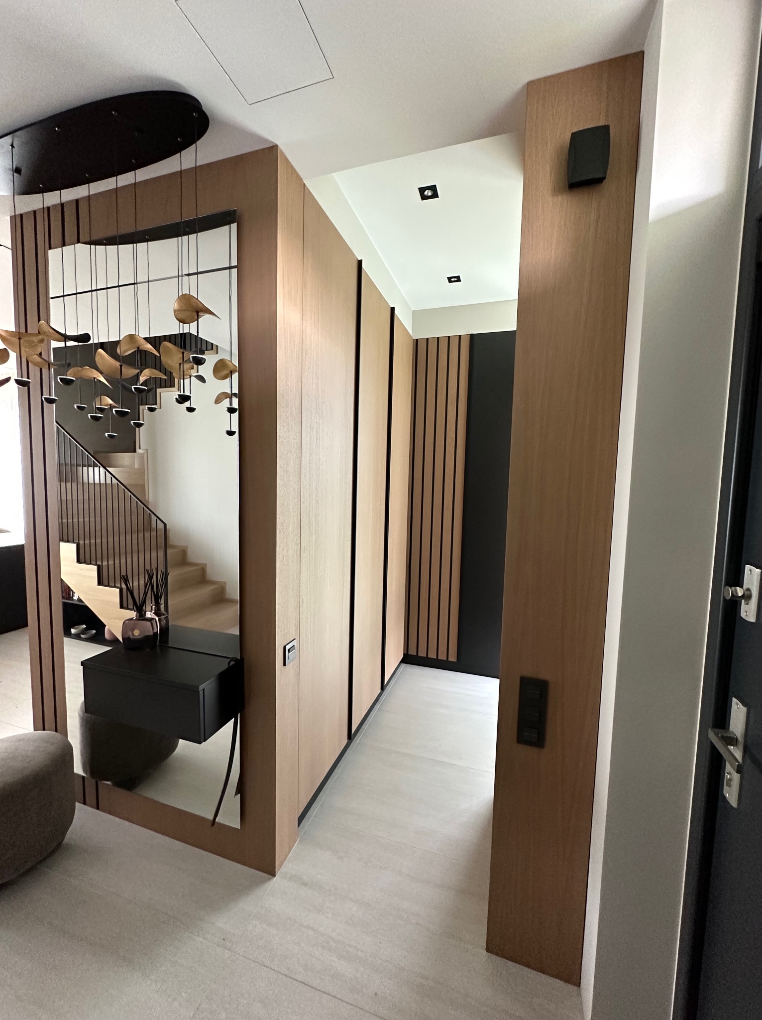 Nowoczesny przedpokój z drewnianą zabudową szafy wnękowej i lustrem odbijającym schody. Minimalistyczny design, ciepłe oświetlenie i eleganckie wykończenie.