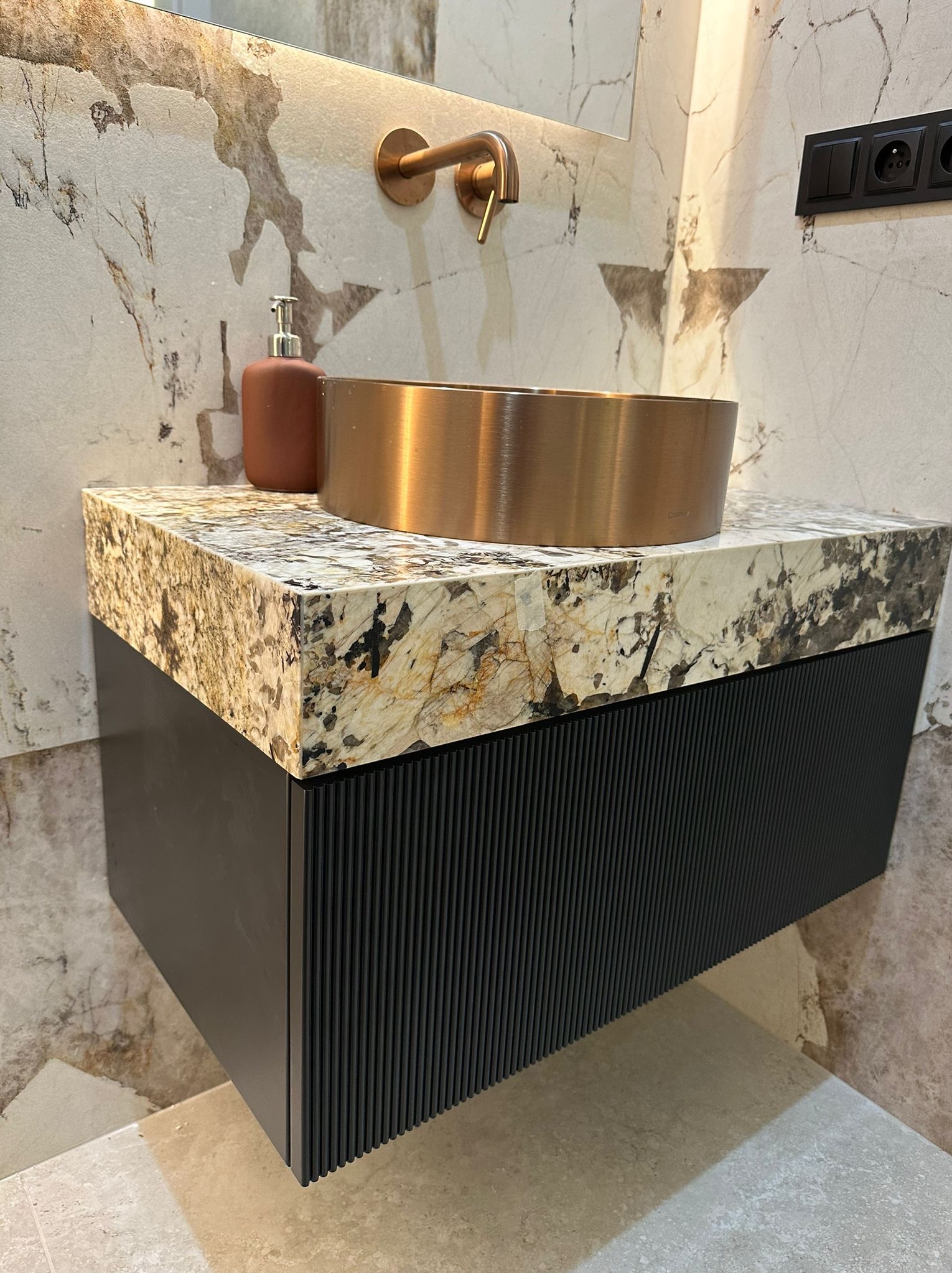 Nowoczesna łazienka: szafka pod umywalkę z blatem z kamienia i ryflowanym frontem, miedziana umywalka i armatura na tle marmurowej ściany.