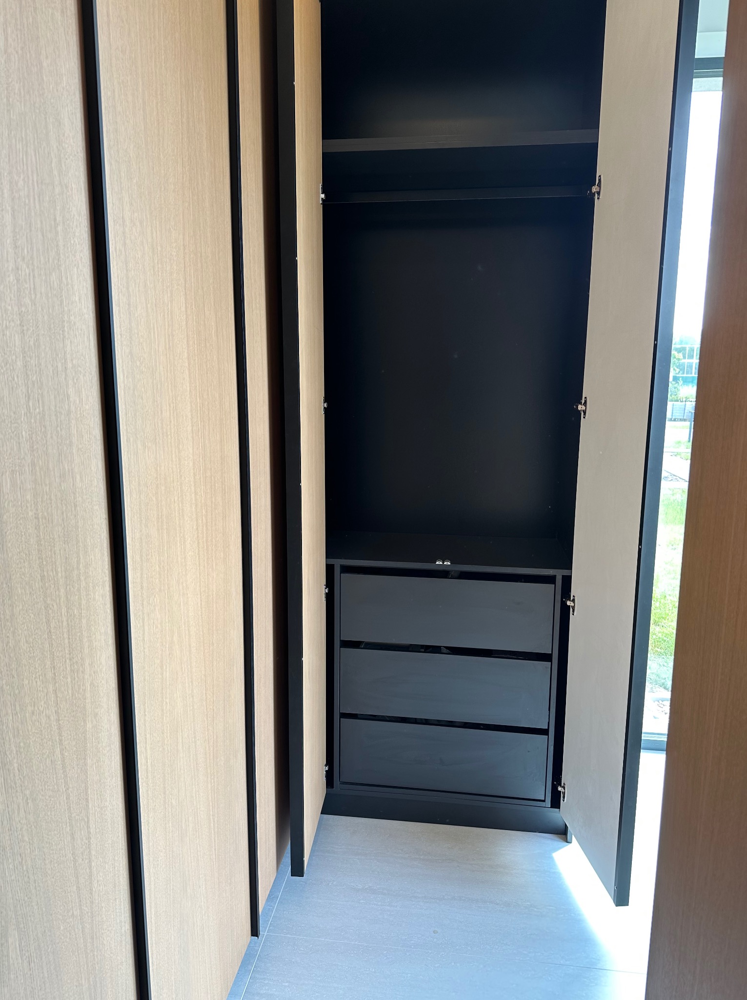 Wnętrze szafy wnękowej z jasnymi frontami i czarnym wnętrzem z trzema szufladami. Drzwi szafy częściowo otwarte, widoczna podłoga z płytek.