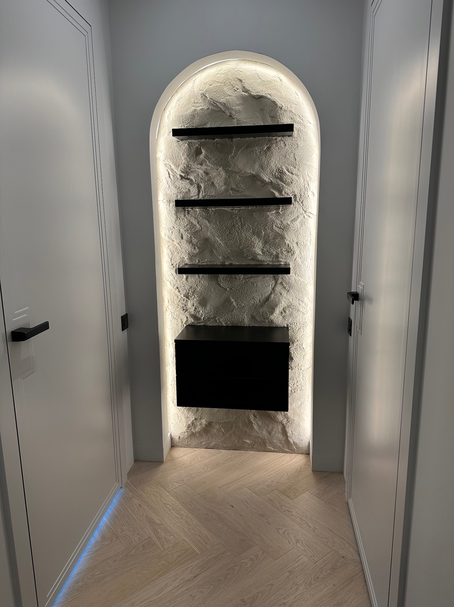 Narożna wnęka z oświetleniem LED, teksturą kamienia i czarnymi półkami, otoczona białymi drzwiami z czarnymi klamkami. Jasny parkiet ułożony w jodełkę.