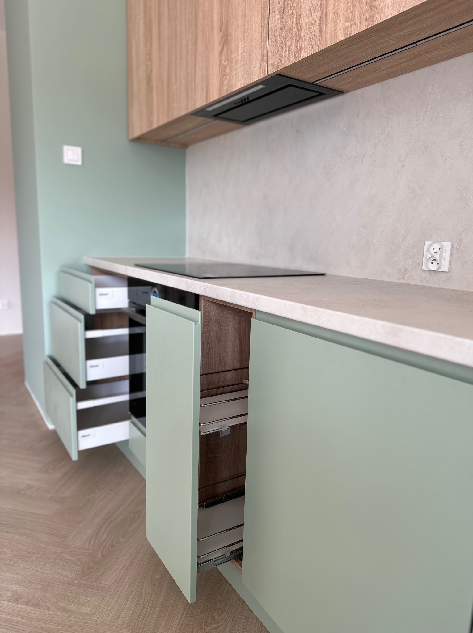Nowoczesna kuchnia z zielonymi szafkami, drewnianymi górnymi szafkami i otwartymi szufladami z organizerami. Widoczna płyta indukcyjna i gniazdko elektryczne.