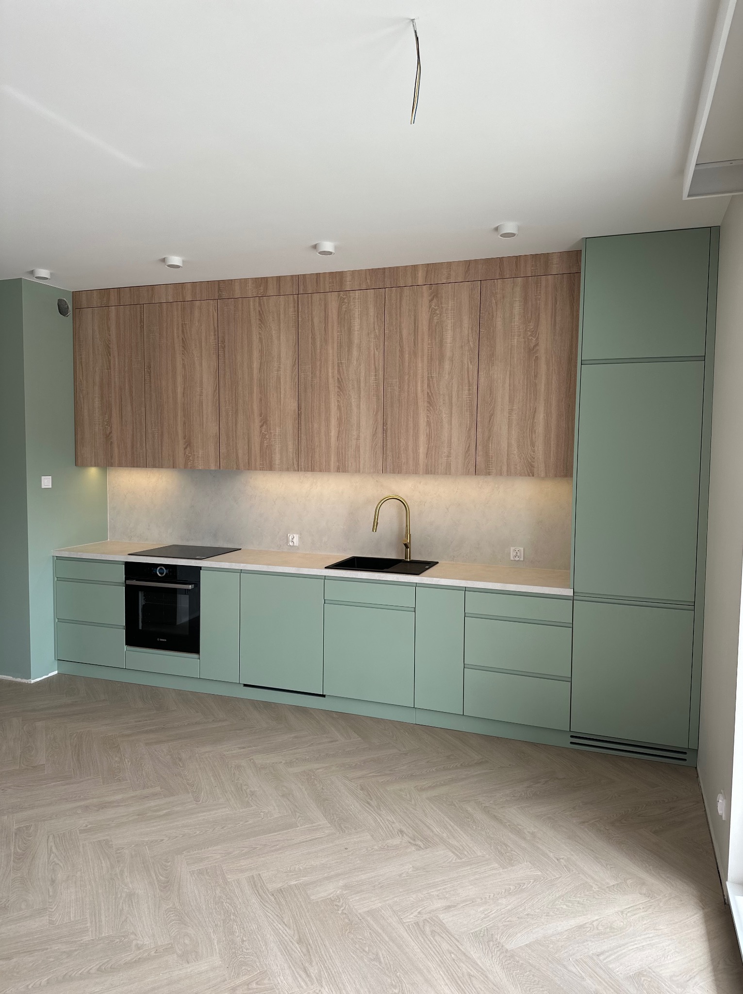 Nowoczesna kuchnia z zielonymi szafkami dolnymi i drewnianymi górnymi. Złota bateria, jasny blat. Na podłodze parkiet ułożony w jodełkę. Minimalistyczny design.