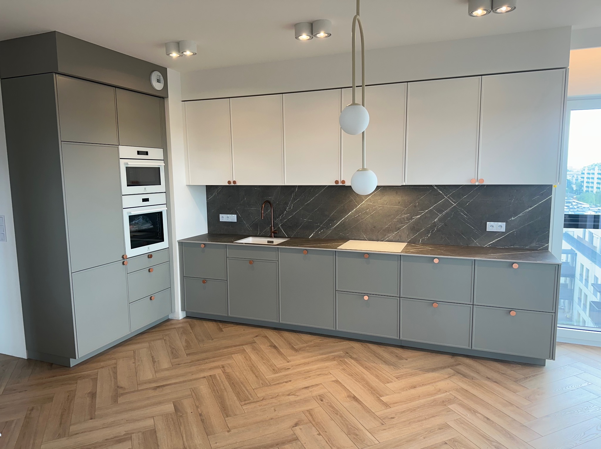 Nowoczesna kuchnia w odcieniach szarości i bieli, z marmurowym blatem, miedzianymi uchwytami i parkietem w jodełkę. Duże okno w tle doświetla przestrzeń.