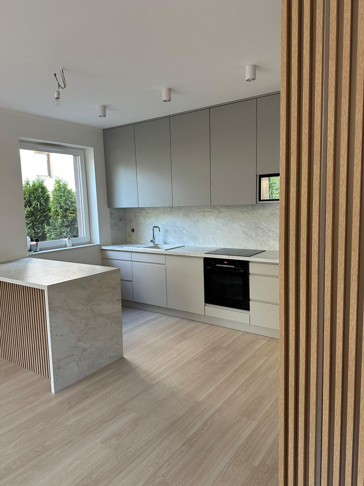 Jasna kuchnia z wyspą, fronty szafek w odcieniach szarości i beżu, marmurowy blat i backsplash. Drewniane lamele w tle dodają ciepła.