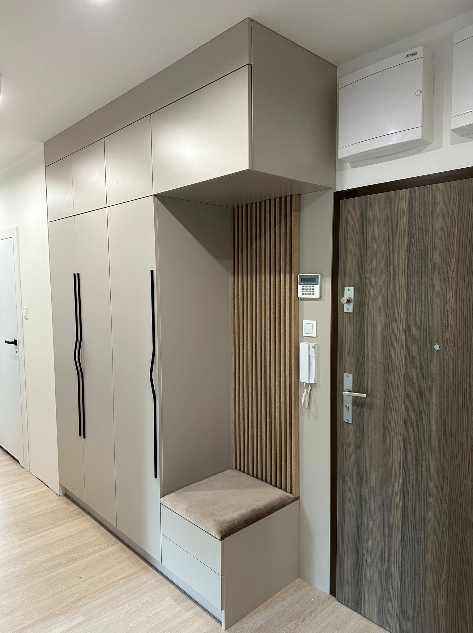 Nowoczesna szafa wnękowa z siedziskiem i pionowymi lamelami w przedpokoju, minimalistyczny design z czarnymi uchwytami i drewnianą okleiną drzwi.