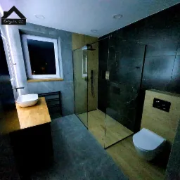 Nowoczesna łazienka z kabiną prysznicową typu walk-in, umywalką nablatową na drewnianym blacie, toaletą podwieszaną i szarą płytką imitującą kamień oraz logo firmy w rogu zdjęcia.