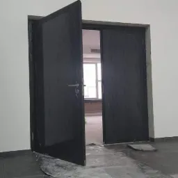 Ciemne, dwuskrzydłowe drzwi wewnętrzne, jedno skrzydło otwarte, widoczne wnętrze pomieszczenia z oknem, szare ściany i podłoga, zabezpieczona folią barierka w rogu.