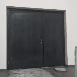 Ciemne, dwuskrzydłowe drzwi z pionowymi żłobieniami i srebrną klamką, zamontowane w białej ścianie, widoczne ślady po ołówku na powierzchni drzwi.