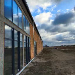 Hala produkcyjna w trakcie montażu rynien i opierzenia, widoczne okna z odbiciem nieba, fragment elewacji z cegły i betonu, oraz teren zewnętrzny z polną drogą.