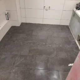 Ułożona, ciemnoszara podłoga z płytek w łazience, z fragmentem dekoracyjnego pasa z geometrycznym wzorem, białe płytki ścienne z zamontowanymi przyłączami wodnymi.