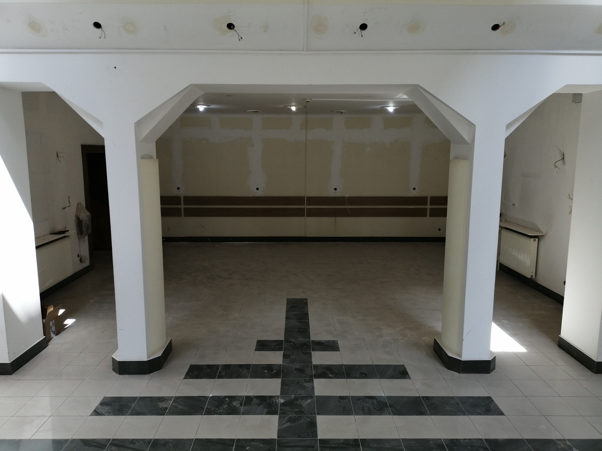 Wnętrze budynku w trakcie remontu, widoczne kolumny, surowy strop z punktami oświetleniowymi bez opraw, podłoga wyłożona płytkami z wzorem krzyża, ściany przygotowane do malowania.