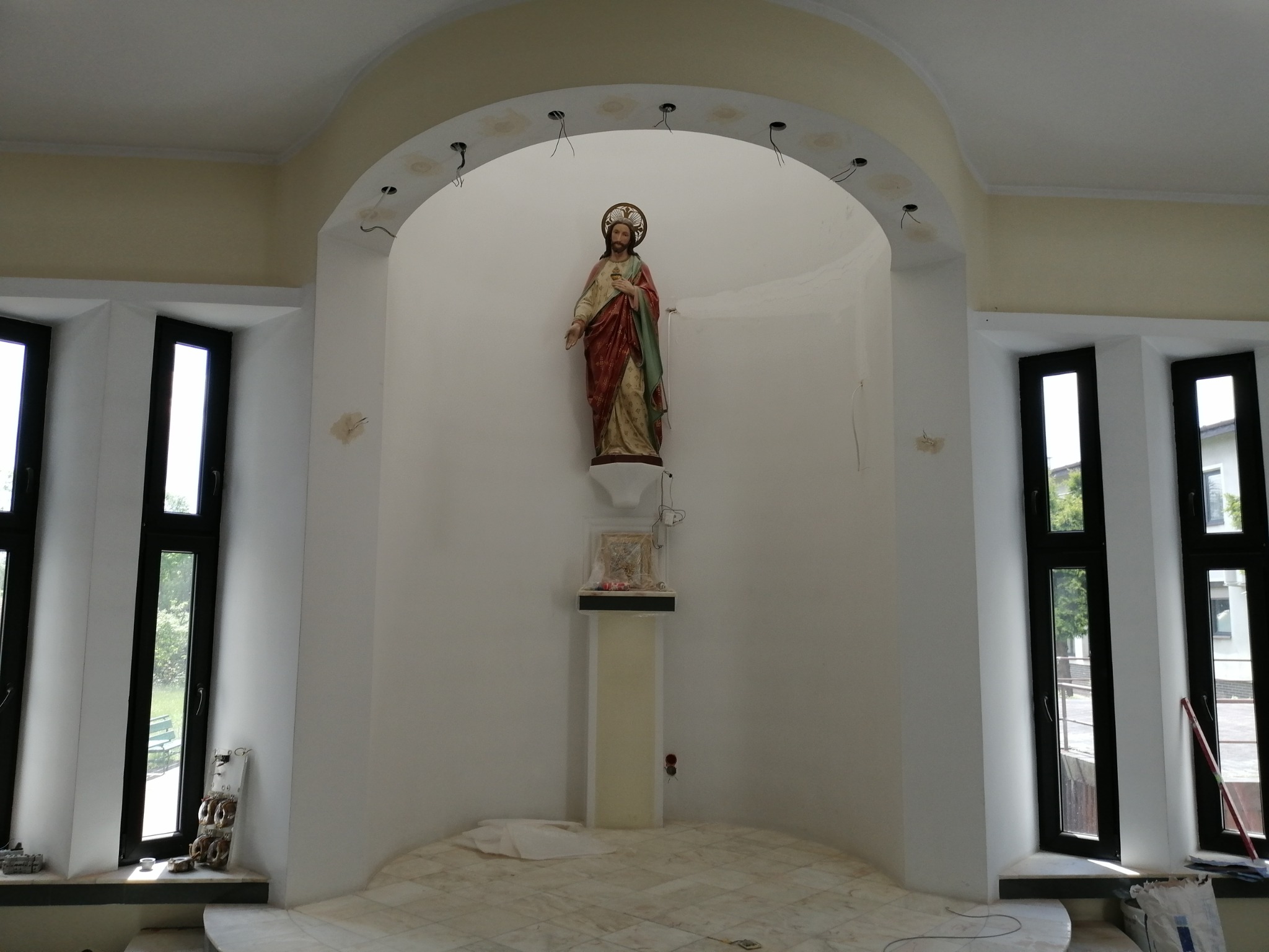 Wnętrze kaplicy w trakcie remontu, widoczne przewody wystające z sufitu, przygotowane pod montaż oświetlenia, oraz statua Jezusa w niszy.
