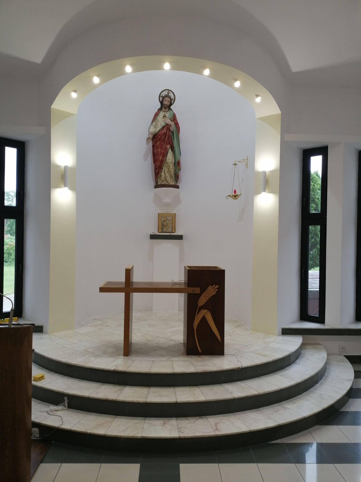 Oświetlenie kościelne: Reflektory punktowe nad łukiem, kinkiety ścienne obok okien, marmurowe schody prowadzące do ołtarza z drewnianą amboną z motywem zboża.