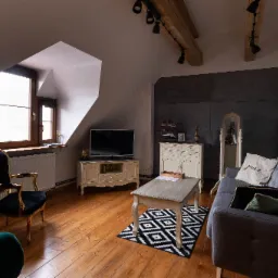 Salon na poddaszu z drewnianą podłogą, szarą sofą, czarnym fotelem z pozłacanymi zdobieniami, geometrycznym dywanem i drewnianymi belkami stropowymi z oświetleniem punktowym.